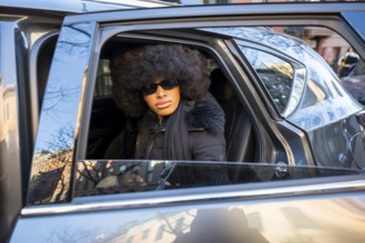 Black woman with a confident expression, wearing sunglasses and a stylish afro, is sitting in a car
