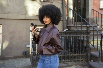Young woman with afro hair wearing a brown jacket and jeans, looking at the camera while holding a