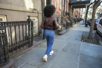 Young woman with afro hair walking along a manhattan sidewalk past brownstones, casual denim jacket