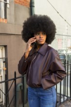 Young african woman with afro hairstyle talking on phone outdoors, wearing a brown jacket and blue