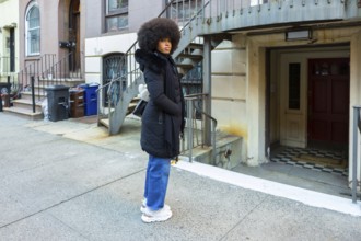 Young woman with large afro hair wearing a black winter coat and blue jeans standing on a city