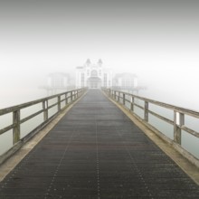 A long wooden walkway leads through thick fog to a white building, view of pier in Sellin in thick