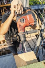 A construction worker wearing a safety helmet uses a drill to drill a hole in a wooden beam,
