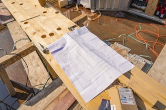 A construction plan is lying on wooden planks on a construction site, surrounded by cables and