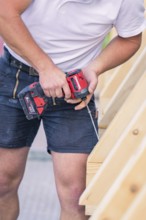 A construction worker in shorts holds a screwdriver and works on a wooden structure, carpentry, new