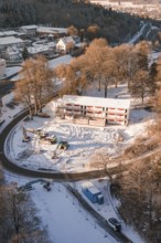 Snow-covered construction site with heavy equipment, surrounded by winter trees seen from the air,