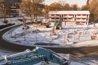 Snow-covered construction site with several excavators and a large building in the background,