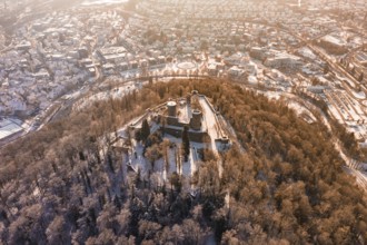 Aerial view of a snowy castle surrounded by a forest, with a city in the background at sunrise,