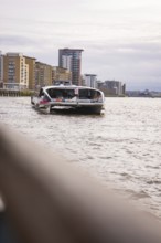 A boat on a wide river with modern buildings in the background and a cloudy sky, London, England,