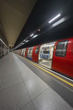 An incoming red subway train stands at an illuminated platform in a dark train station, London,