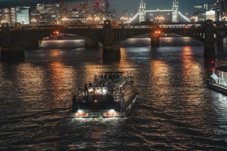 Boat under an illuminated bridge on a river at night, city lights reflecting in water, London,