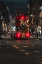 Double-decker bus travels at night through an illuminated city street surrounded by buildings,