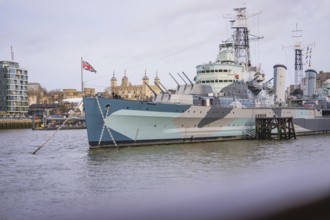 Historic warship on the riverbank in a city with British flag and buildings in the background, HMS