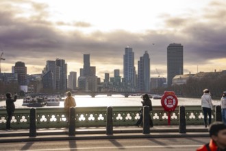 View across a bridge to the skyline of a city with river and cloudy sky, people in the foreground,