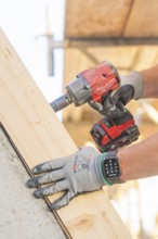 A worker with gloves uses a cordless screwdriver on a large piece of wood, carpentry construction,