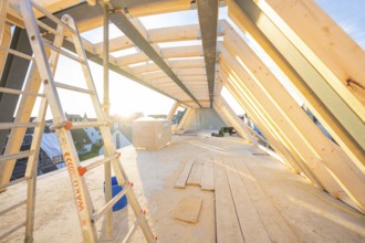 An unfinished wooden roof truss on a construction site, illuminated by sunlight. A ladder is ready,