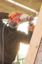 A worker uses a drill to process a slanted wooden board under sunlight, carpentry construction,