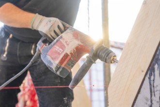 A worker with a red-black drill works on a wooden plank in sunlight, carpentry construction, roof