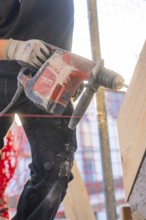 A worker is drilling wood with a red electric drill on a construction site. Dust flies up,