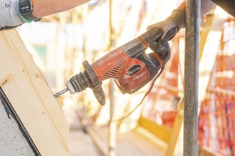 A worker drills into wood with a red drill while sunlight falls through the construction site,