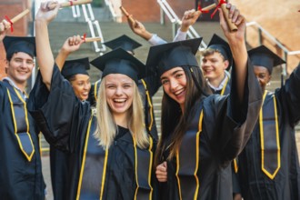 Group of happy student graduates wearing caps and gowns, cheering and holding rolled up diplomas,