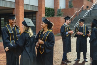 Happy diverse students wearing graduation caps and gowns, holding diplomas, and conversing outdoors