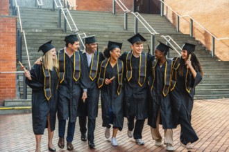 Diverse young college students walking together in graduation gowns and caps, holding diplomas,