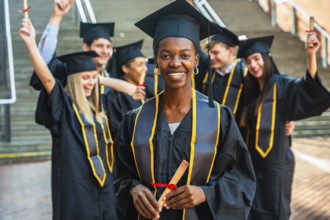Diverse group of smiling young graduates in caps and gowns on campus, holding diplomas and