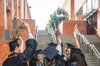 Group of diverse graduating students wearing academic caps and gowns, happily raising their diploma