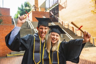 Young man and woman friends wearing graduation caps and gowns, cheering and holding diplomas