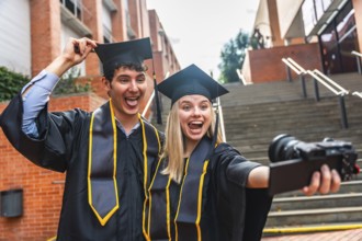 Two cheerful university graduates, a man and a woman, wearing cap and gown, smiling and making