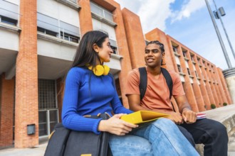 Two young diverse students are sitting on a wall outside a university building, engaging in a