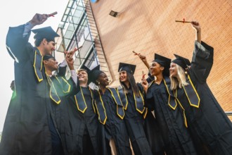 Diverse group of joyful young graduates in black caps and gowns celebrating on campus, holding