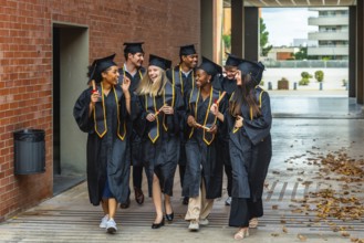 Diverse group of happy graduates celebrating achievement, walking on campus wearing academic gowns,
