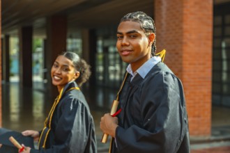 Two diverse university graduates smile and pose on campus in caps and gowns, proudly holding
