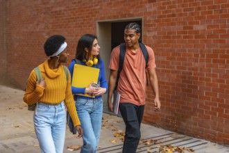 Three diverse young university students walking along a brick wall, carrying backpacks and books,