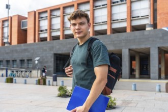 Young man student carrying a backpack and blue notebook, confidently walking on a modern university