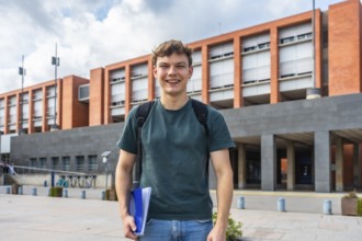 Young male student with backpack and folders standing outdoors on campus, smiling confidently at