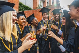 Young diverse international students wearing graduation caps and gowns toasting with champagne