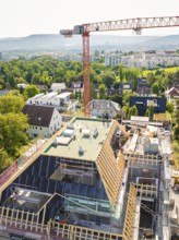 A construction site with a crane over a roof, an urban and green landscape in the background,