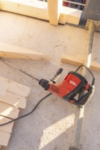 A red drill is lying on a concrete floor between wooden planks in bright surroundings, carpentry