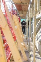 A worker walks over scaffolding surrounded by wooden boards and a protective net in a bright