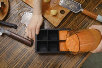 A person is pouring fruit mixture from a blender into a silicone ice cube tray. The table has