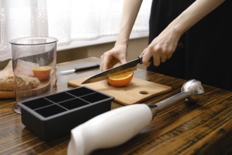 A person is cutting an orange on a wooden board with a knife. There are other kitchen items nearby,