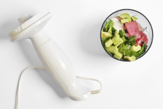 A white hand blender rests beside a bowl filled with chopped tuna, avocados, and herbs. The process