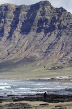 Beach with rocks and cliffs (Risco de Famara) in the background, people enjoying the sea, Caleta de