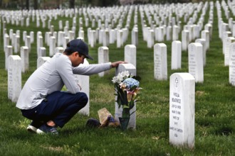A man kneels in front of a tombstone in a large cemetery and deposits a bouquet of flowers,