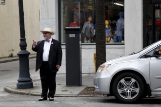 Elderly man in formal clothes stands in front of a shop window on a street corner. Modern car