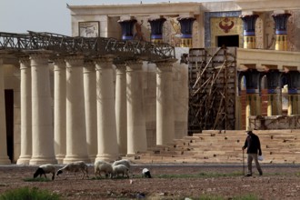 Film set with ancient columns and decoratively decorated building, a human leading goats past,