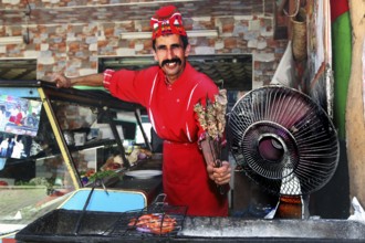 Smiling chef in red outfit offers brochettes at a barbecue area in Moulay Idris, Moulay Idris,
